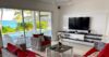 Living room in a St Barts villa with rattan seating, red cushions, wall-mounted TV, and sliding glass doors to pool and sea views