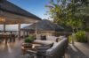 a modern outdoor lounge area with a gray sectional sofa and pillows on a wooden deck, with a view of the dining area and the surrounding tropical plants