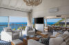 a spacious living room with white sofas and a view of an outdoor dining table on the terrace, with the ocean in the background