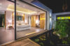 The interior of a contemporary bedroom with a large circular bed. The room has a glass wall that separates it from a tranquil, black-bottom pond. Beyond the pond, a wooden deck with white furniture and a view of the ocean is visible.