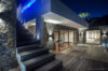 A night shot of a wooden deck with an infinity pool. The deck has a row of white sun loungers and a separate lounge area with glowing, color-changing outdoor sofas and a coffee table, all illuminated against the dark night.