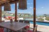 Covered outdoor dining terrace with wooden table, woven pendant lamps, and lagoon views by the pool at a St Barts villa.
