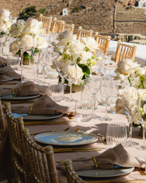 Elegant outdoor dining table with light blue-grey plates with gold patterns, gold cutlery, white rose centerpieces, and scenic hillside backdrop at St. Barts villa.