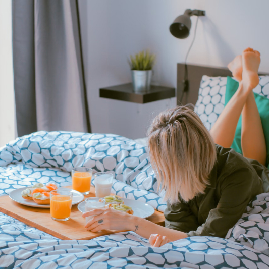 Relaxed breakfast in bed with woman holding milk glass, fruit-topped toasts, and orange juice on wooden serving tray.