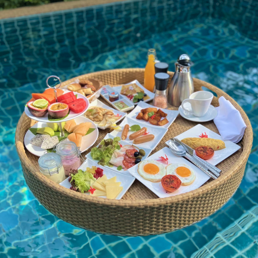 Floating pool breakfast with tropical fruits, pastries, and morning dishes on wicker tray.