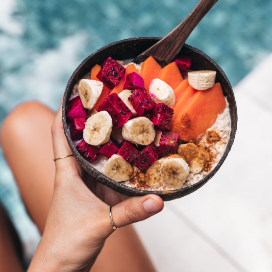 Natural coconut bowl containing dragon fruit, papaya, banana, and grain base with wooden spoon at poolside setting