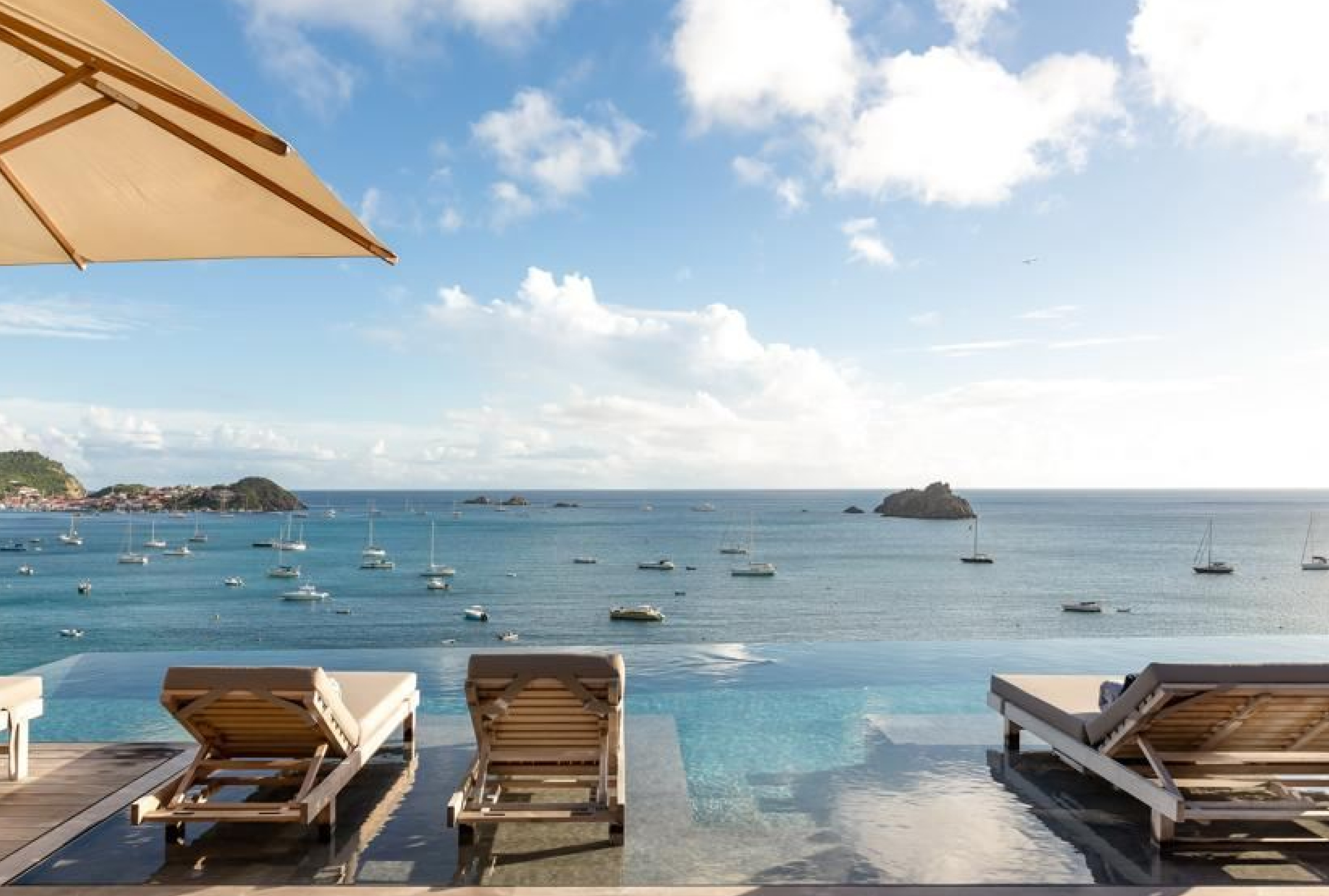 Villa infinity pool with lounge chairs, a bay with boats, and a coastal town in Corossol, St. Barts