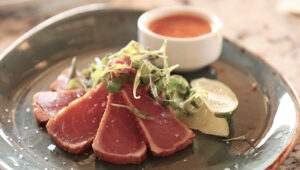Seared tuna slices with greens and dipping sauce on a ceramic plate.