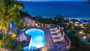 Lit pool and lounge chairs on a deck overlooking the ocean at dusk