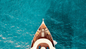 Aerial view of a yacht’s bow sailing on clear turquoise ocean water.