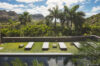 Sunny poolside lawn with modern sun loungers surrounded by palm trees and mountain views under a bright blue sky.