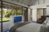 Bright bedroom with a plush bed, pop-up TV, and glass doors opening to a lush tropical garden and wooden deck.
