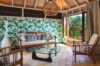 Bright tropical living room with botanical wallpaper, wooden furniture, and large windows opening to lush greenery.