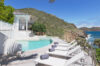 Cliffside infinity pool with white loungers overlooking the ocean and green hills under a clear blue sky.