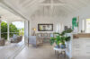 Bright open living space with white vaulted ceilings, cozy seating, and tropical plants leading to a terrace with ocean views.
