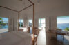 Bright bedroom with a four-poster bed and open doors revealing panoramic ocean and hillside views